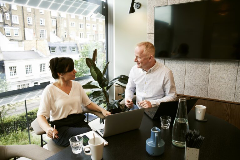 two people sit at a desk facing each other in a sun-lit office with a computer monitor in front of them a TV on the wall behind them.