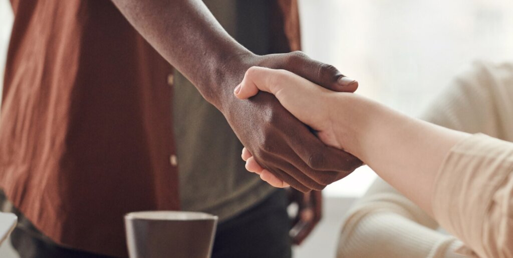 two people shake hands across a conference table where one person is standing and one person is seated